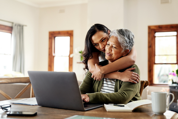 Young girl hugging grandmother who is using laptop.